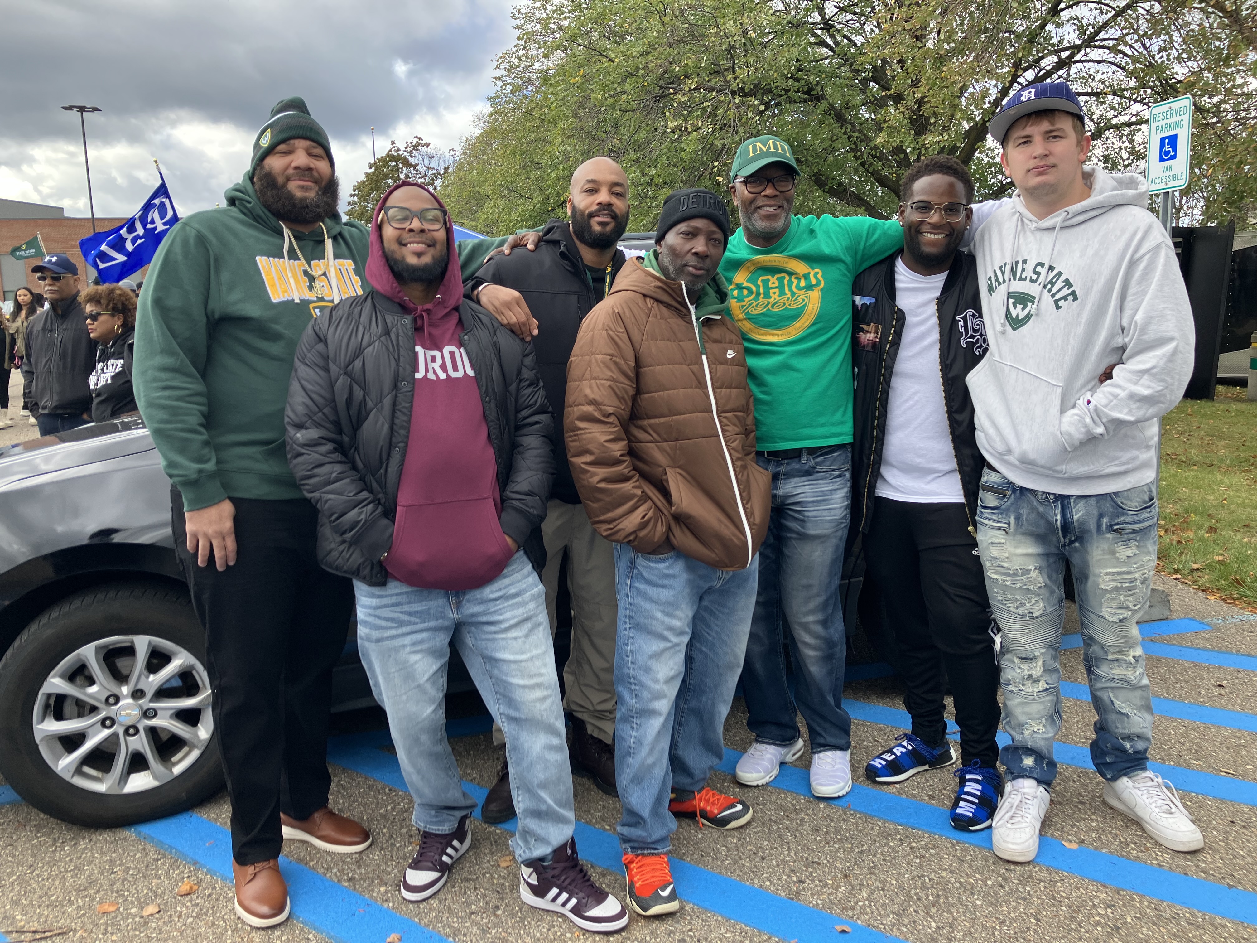 Seven men standing together in front of a car