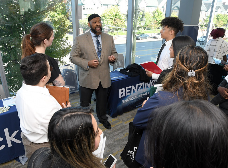 A man speaking in a grey suit, surrounded by students listening in. 