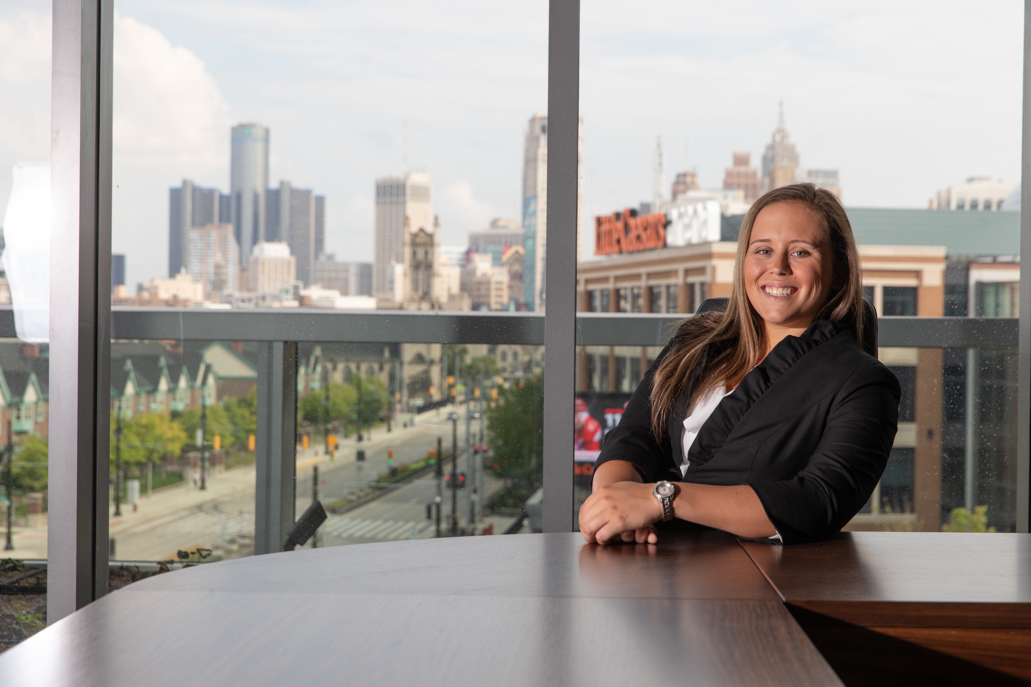 A female business student sits at a table smiling in front of a conference room window showing downtown Detroit