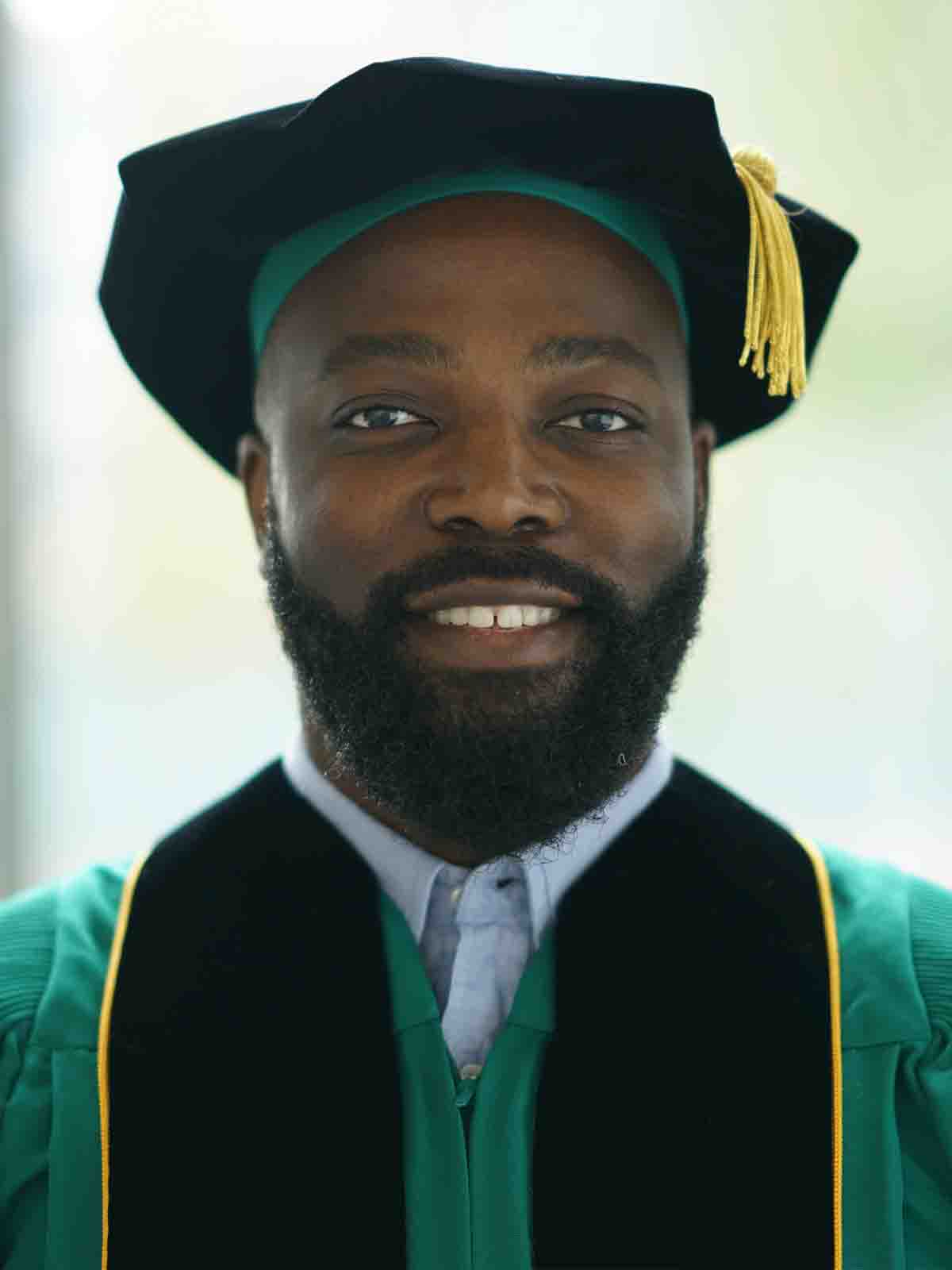 A male doctoral business student smiles for a headshot while wearing his graduation cap and gown