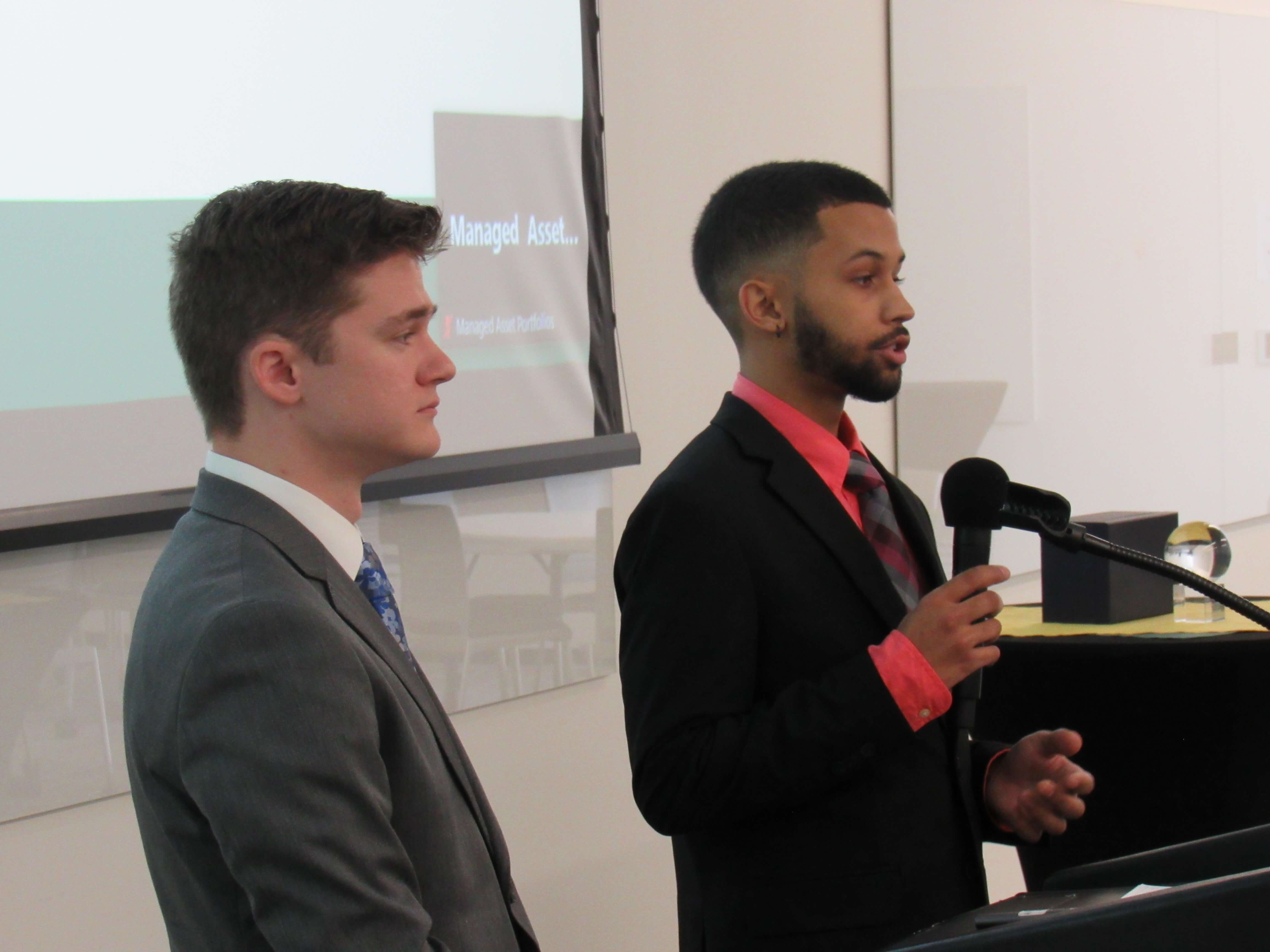 Two male finance students in suits stand in front of a projection screen presenting during the 2023 Stock Pitch Competition