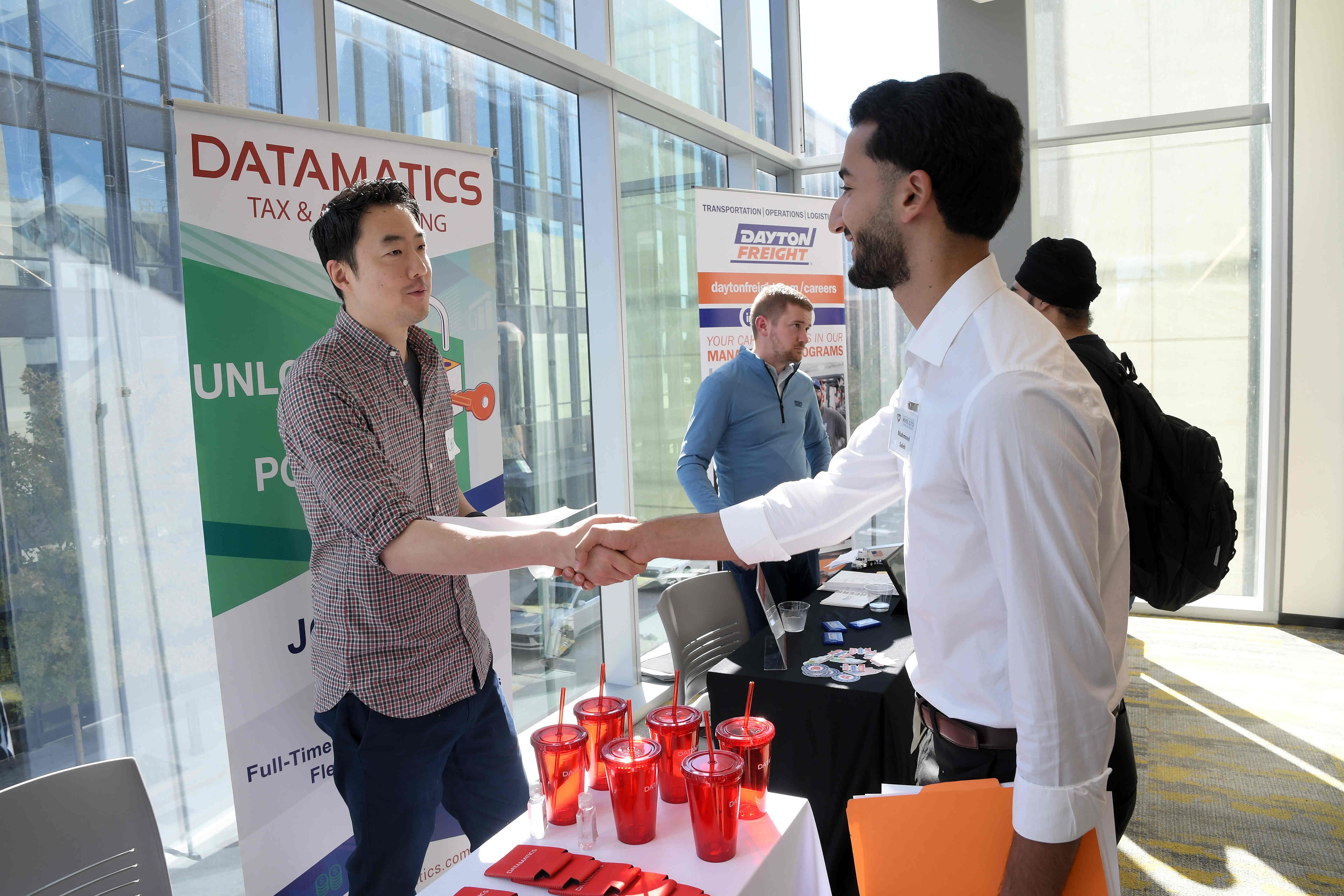 Two men shake hands over a table with six red cups on it. 