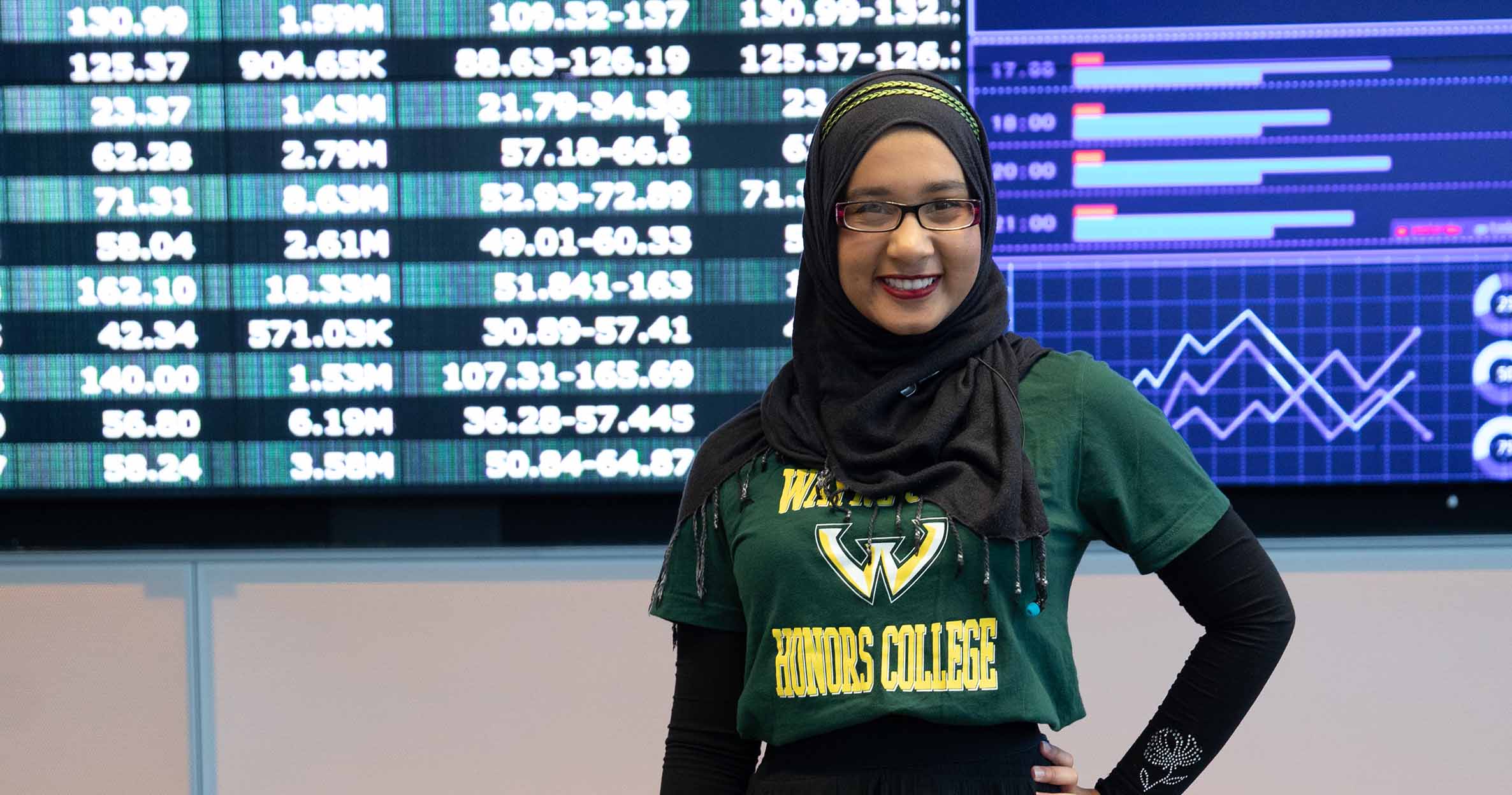 A female business students stands with a hand on her hip, smiling in front of a screen in Ilitch School&#39;s Data Analytics Lab