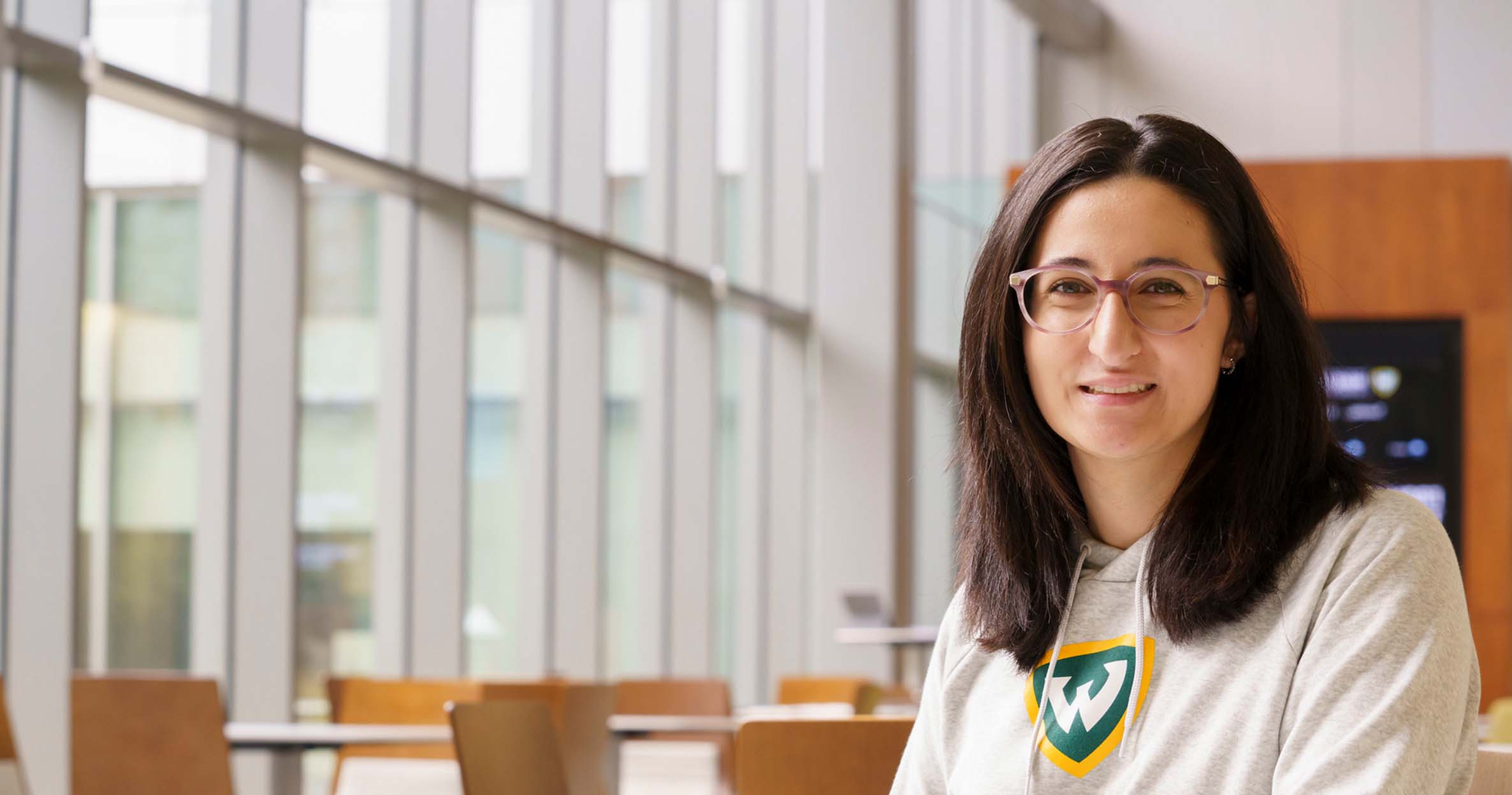 A female business student sits at a table in the Ilitch School atrium wearing a WSU shirt and smiling for a headshot