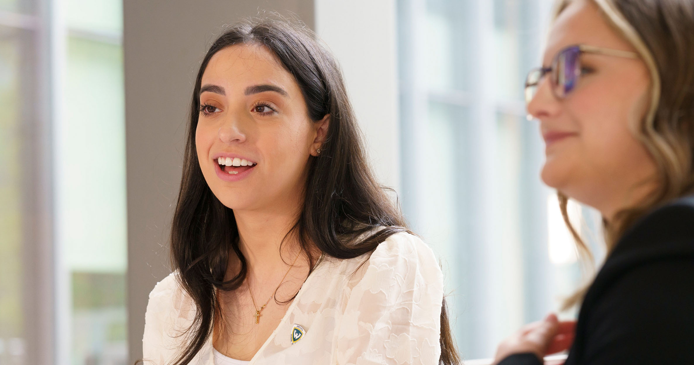 Two female business students sitting in the atrium look to their right, smiling and talking