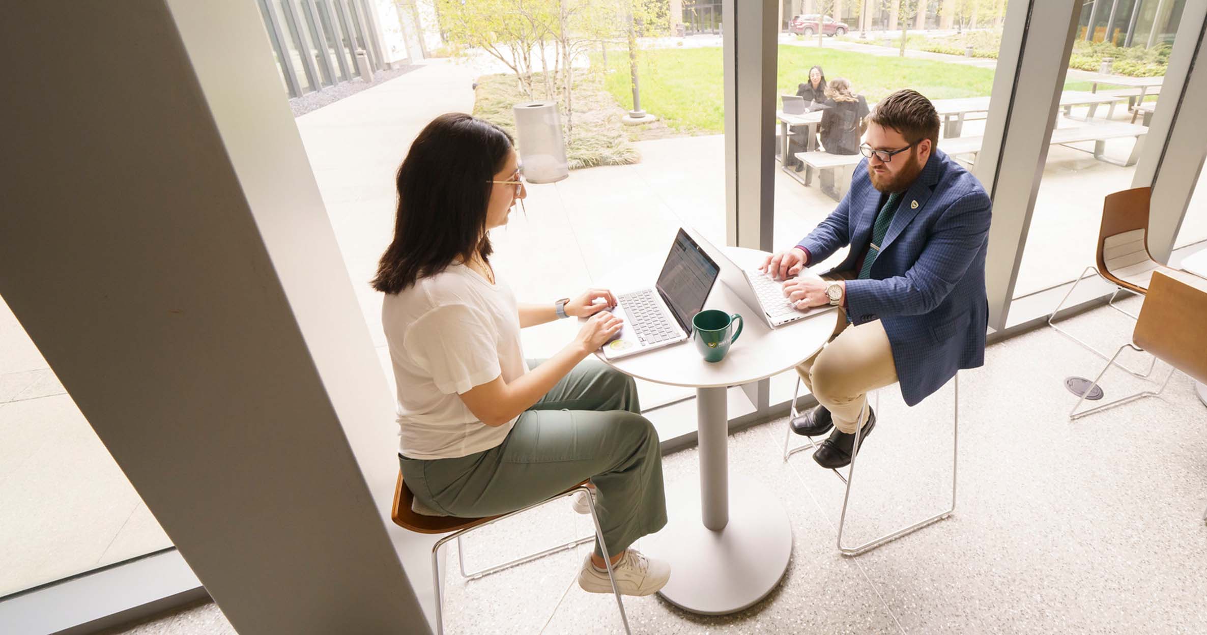 A male student and female student sit across from each other at a table in front of a window typing on their laptops