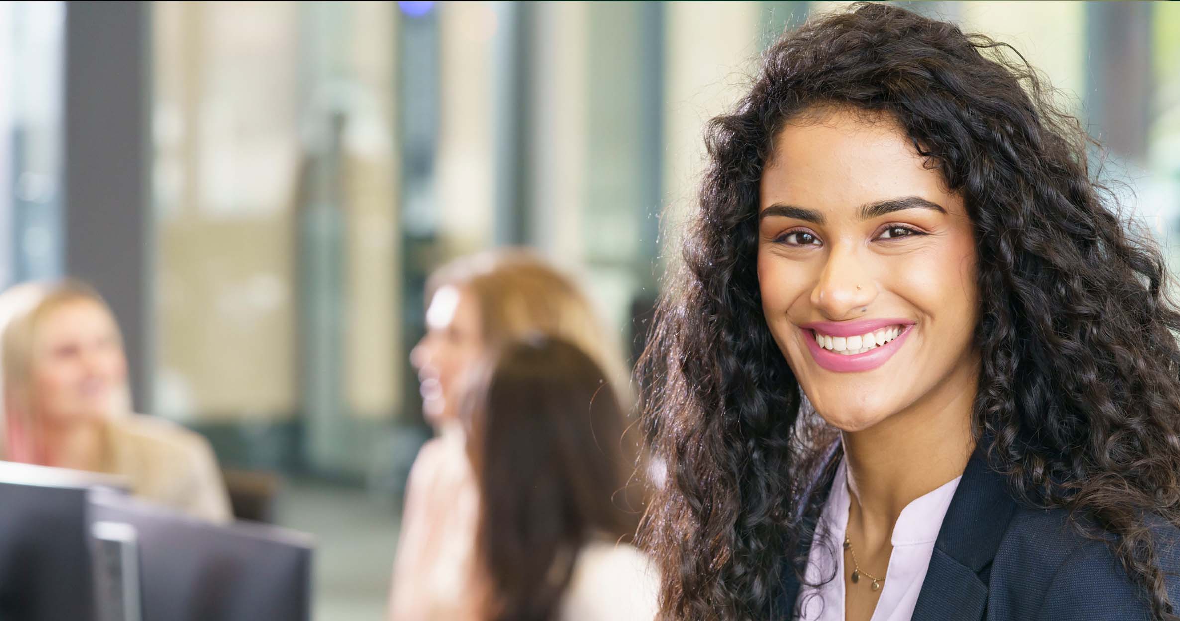 A student with long curly hair wearing a blazer and button down shirt smiles. Three students sit nearby at computers