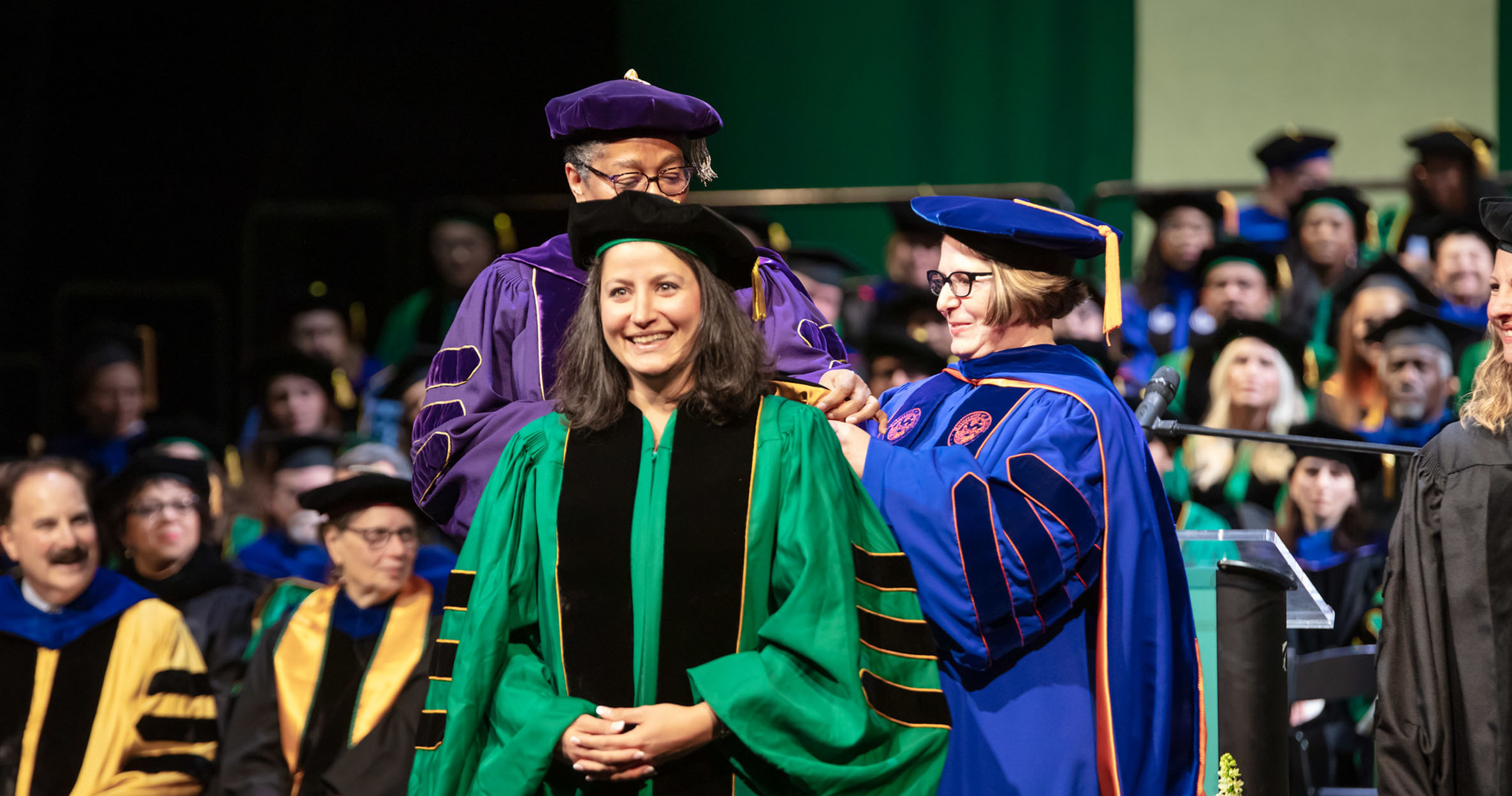 An Ilitch School Ph.D. student wearing a green academic robe stands on stage as faculty present her with a stole