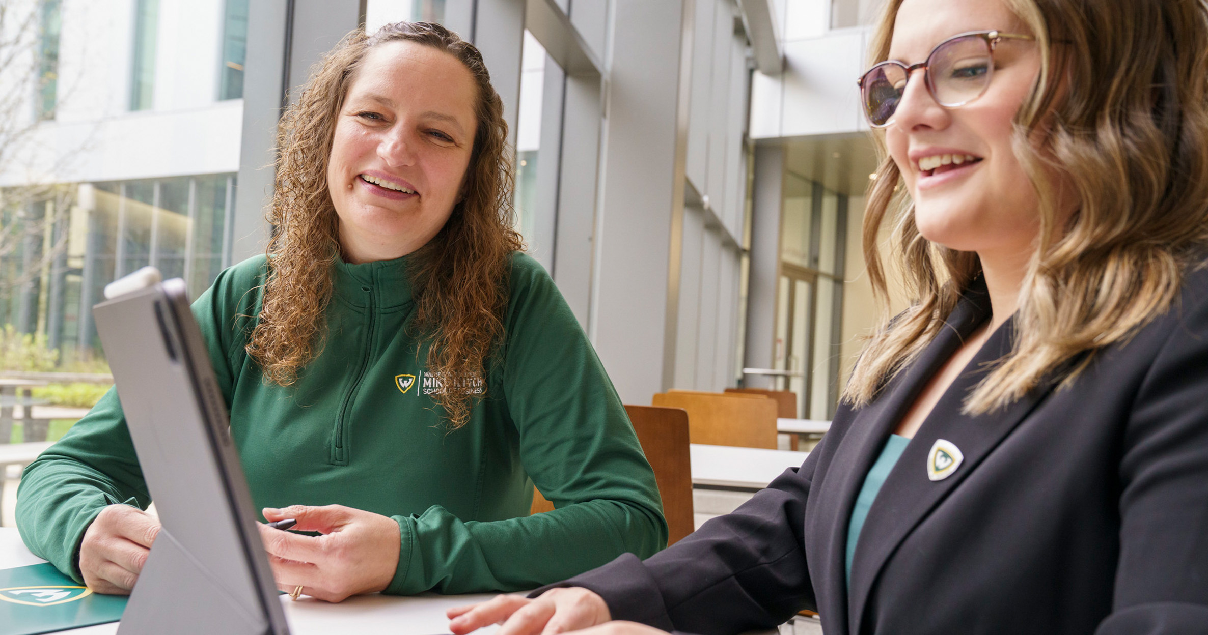 A female business student sits at a table with an Ilitch School staff member, smiling and typing on her laptop  