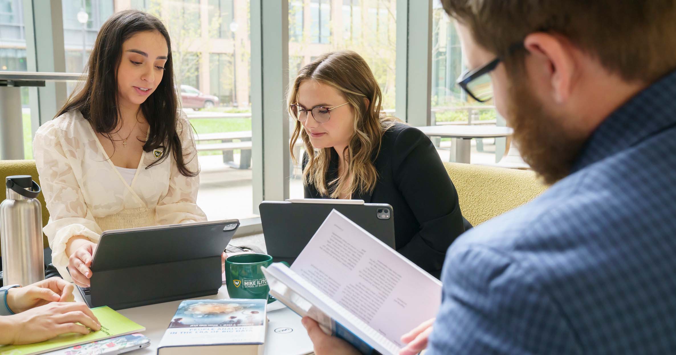 A group of Ilitch School Business students sit around a table in the atrium with their laptops, smiling and talking