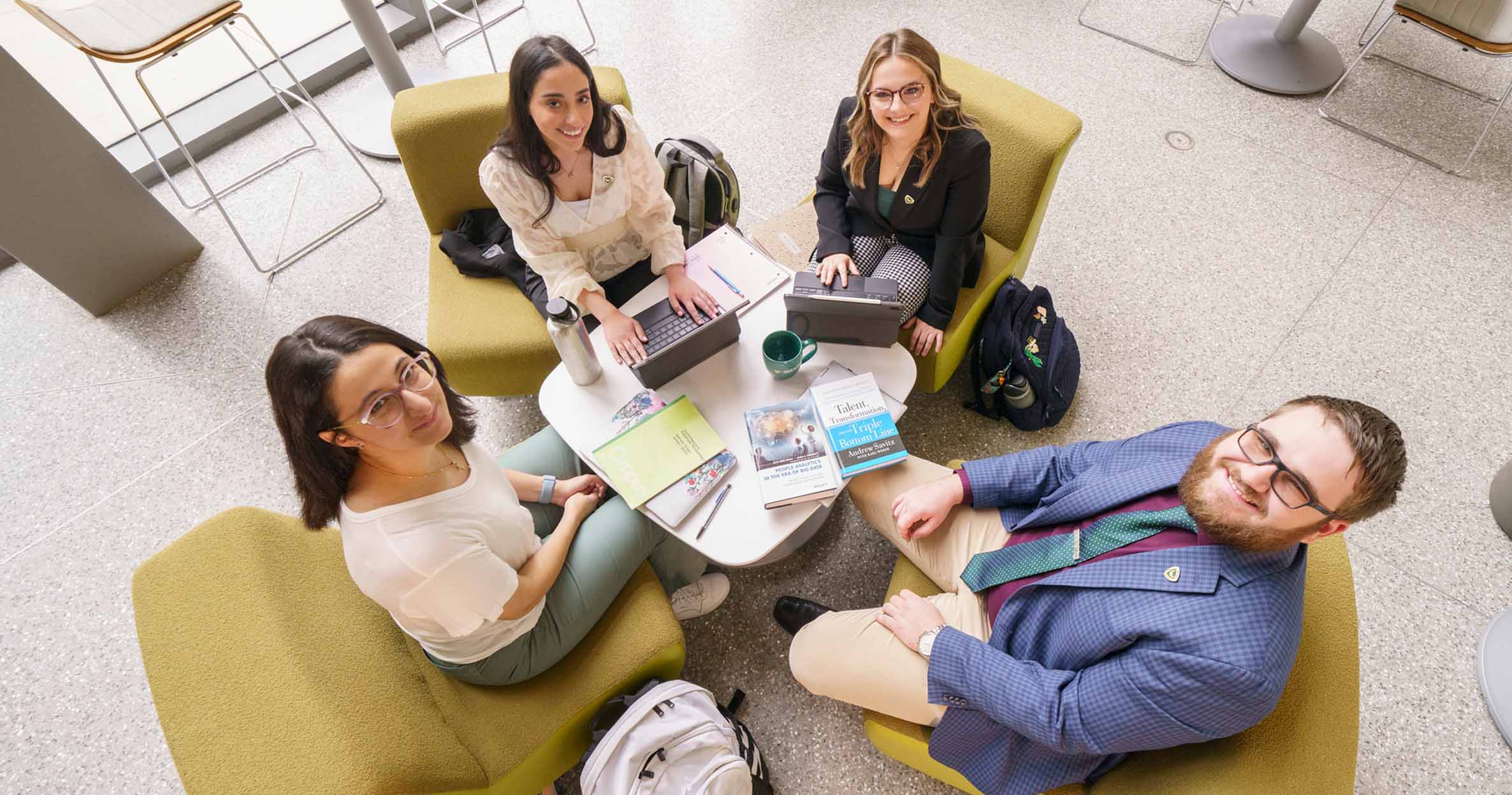 Four business students sitting in green chairs with their laptops in Ilitch School&#039;s atrium look up and smile