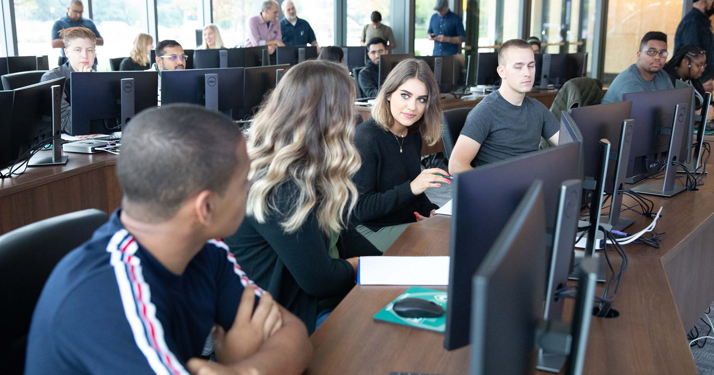 A group of business students sit at a long table with desktop computers in the Data Analytics Lab