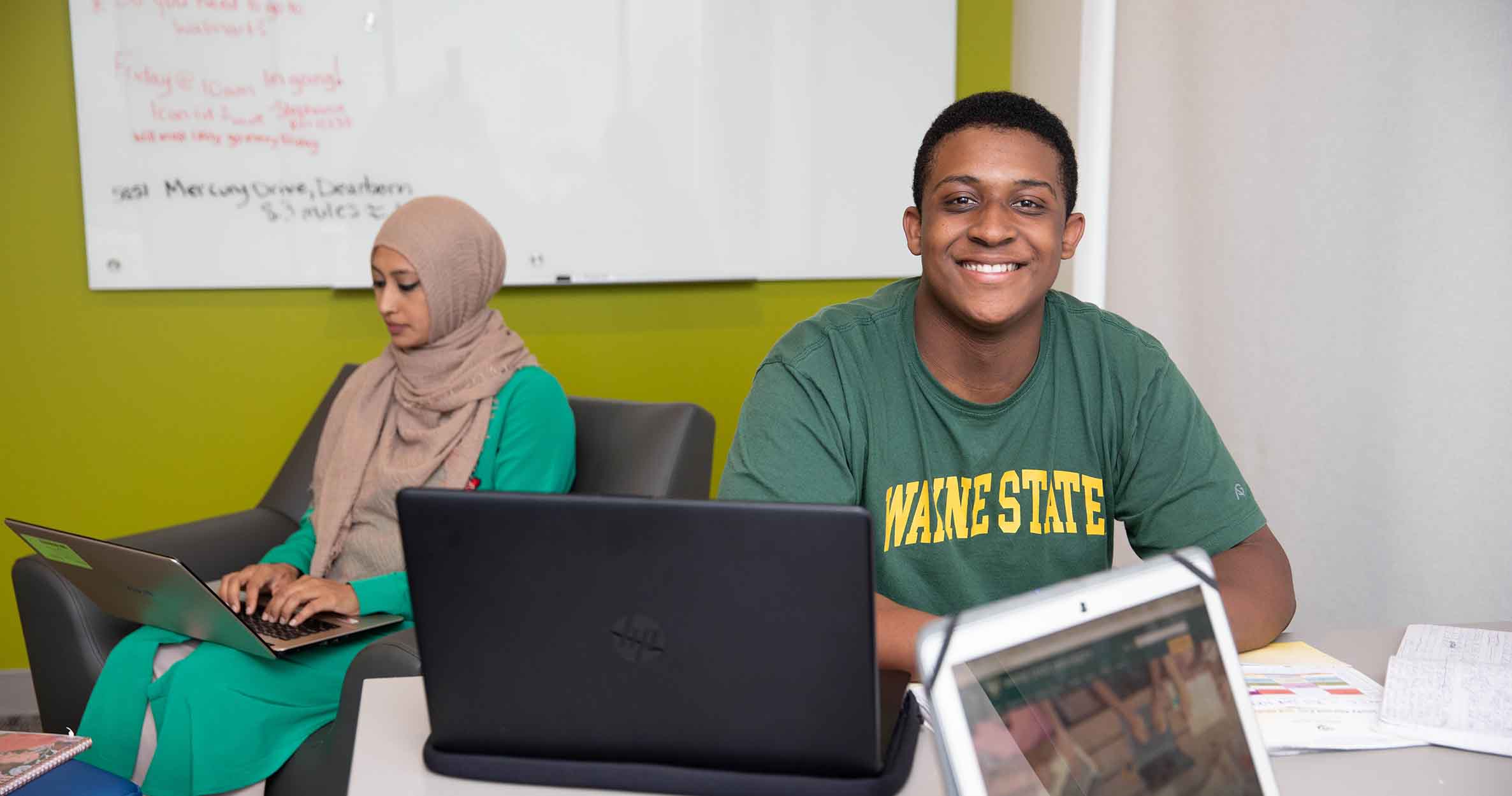 A male business student sits at a table with a laptop smiling and a female student sits beside him with her laptop in her lap