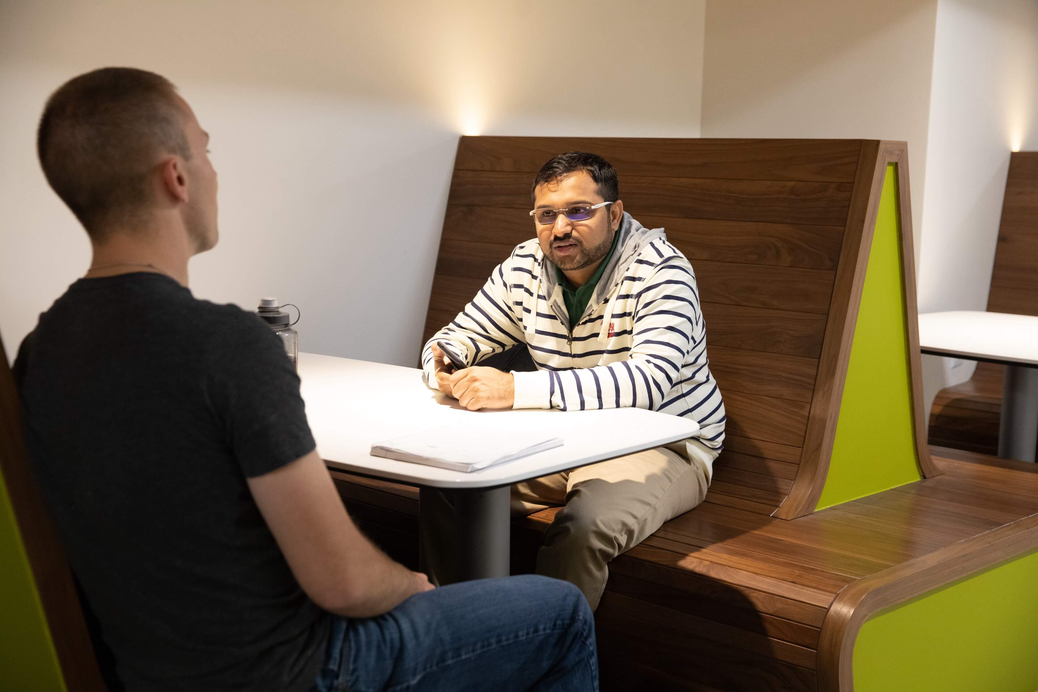 A male is seated on a booth at a table inside talking with another male seated on the opposite side of the table