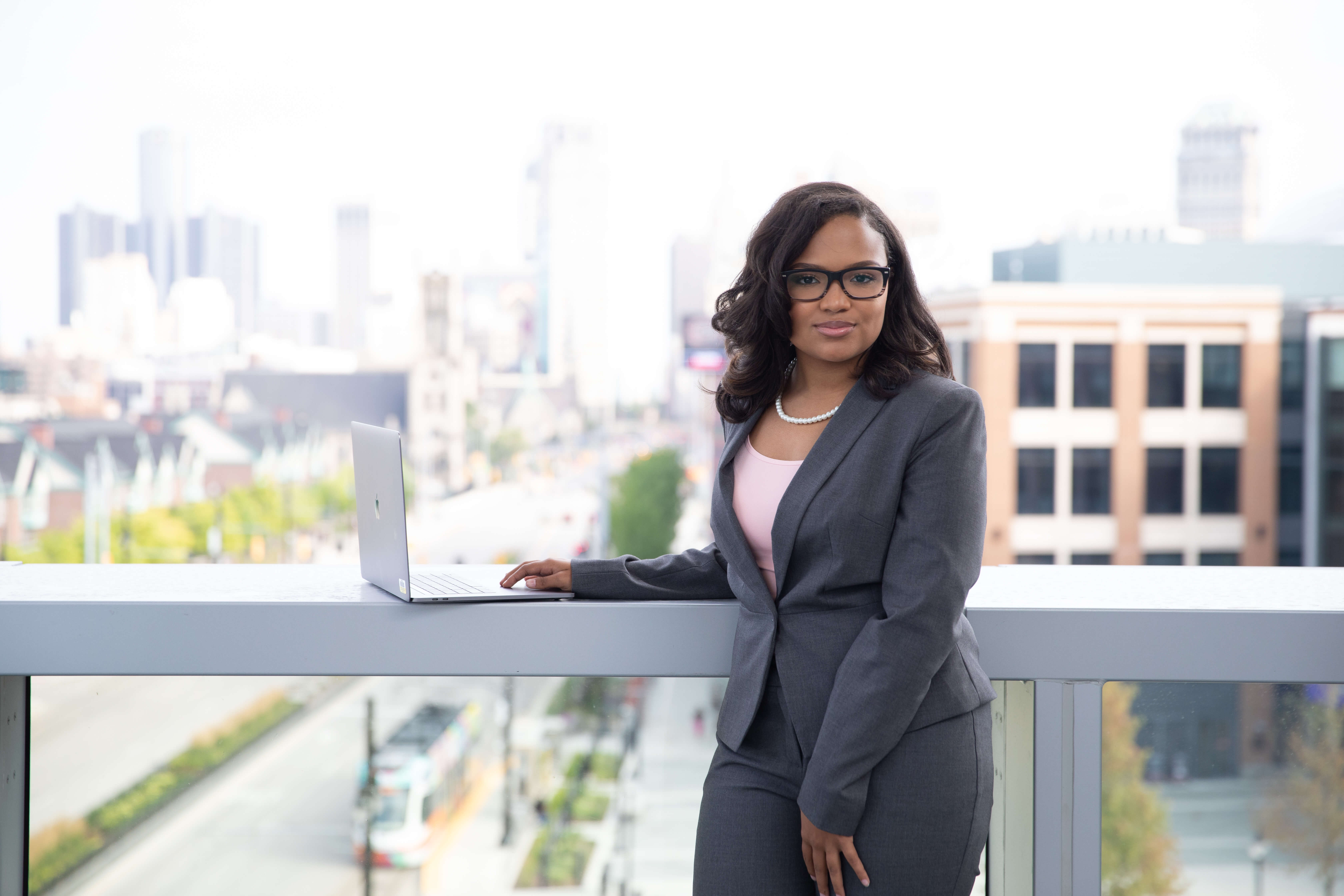 A female business student in a suit stands at a terrace with her hand on her laptop, posing for a headshot