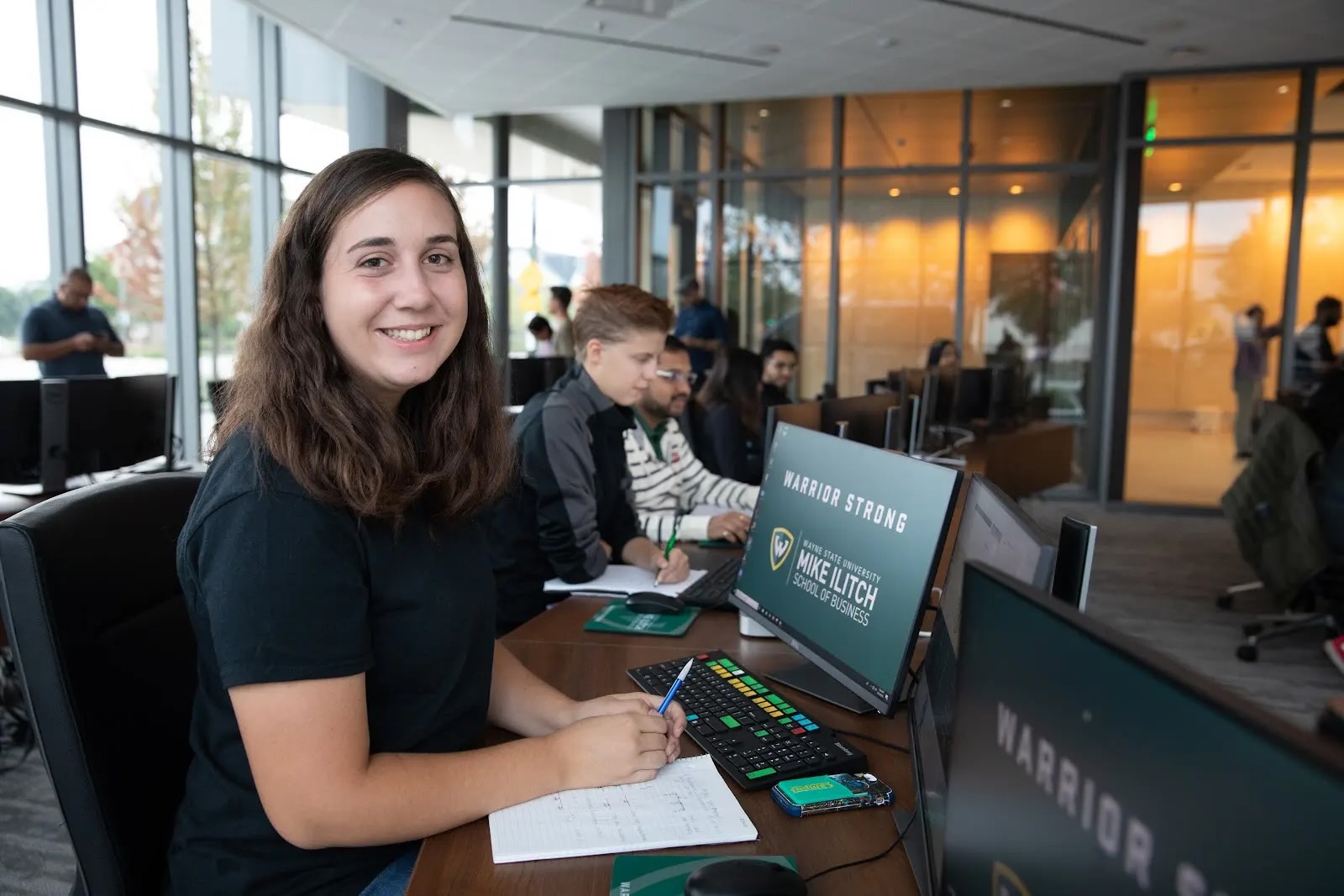 A female business student smiles, sitting at a table in front of a desktop computer with other students working at computers