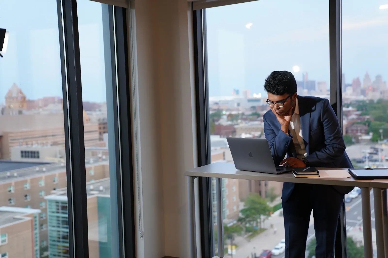 A male business student wearing a suit stands with his laptop in front of a window