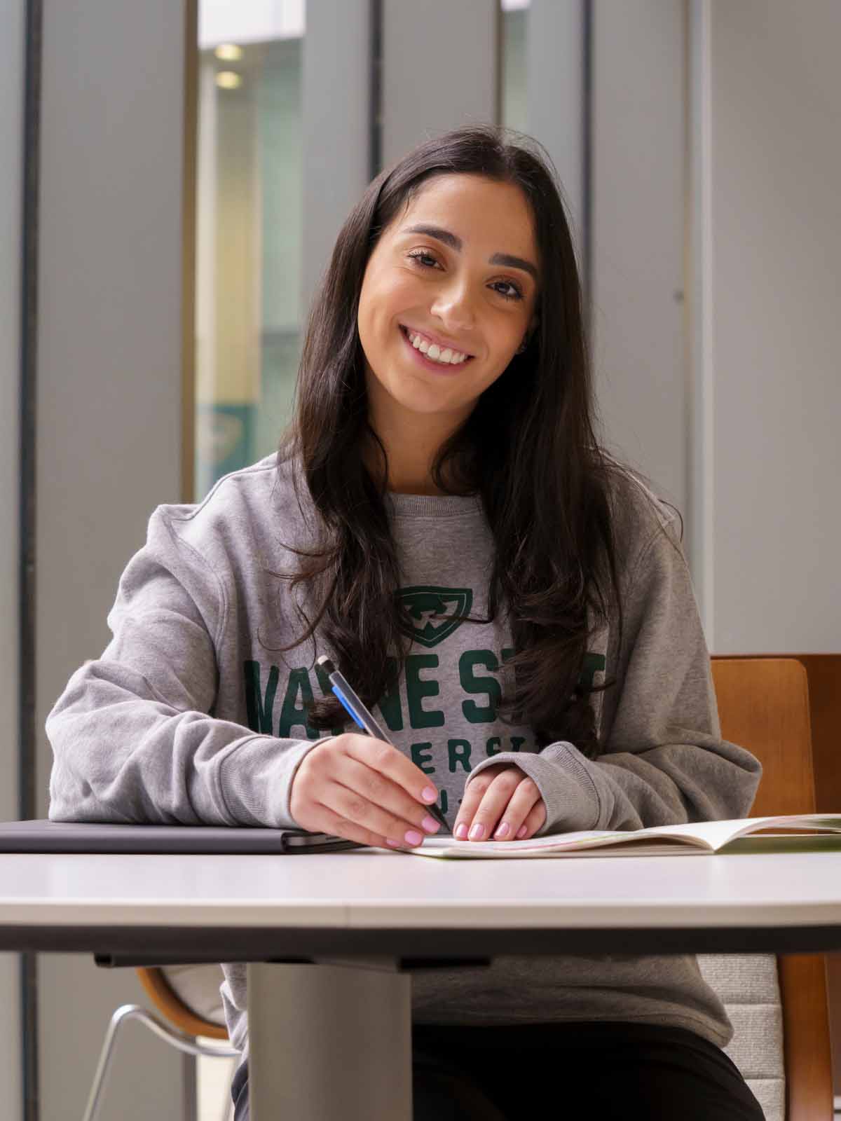 A female business student sits at a table inside an Ilitch School building, looking up and smiling with a pencil in her hand