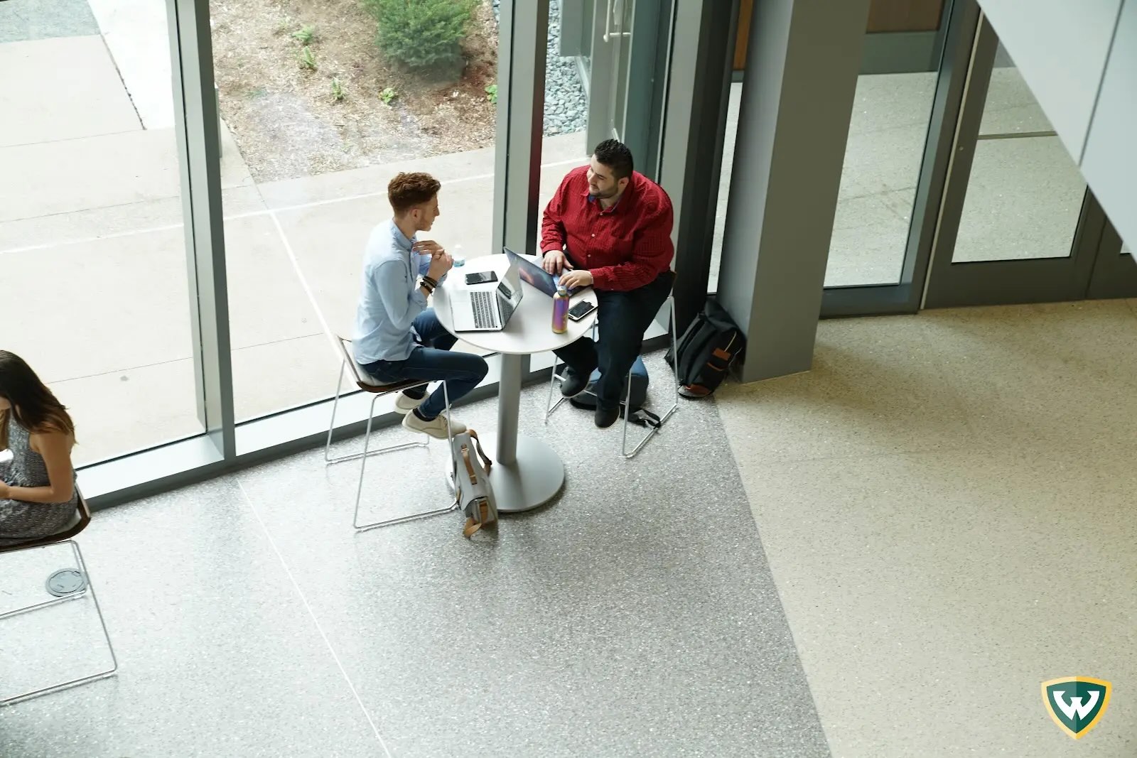 Two males sit at a hightop table in the atrium using their laptops and talking with each other