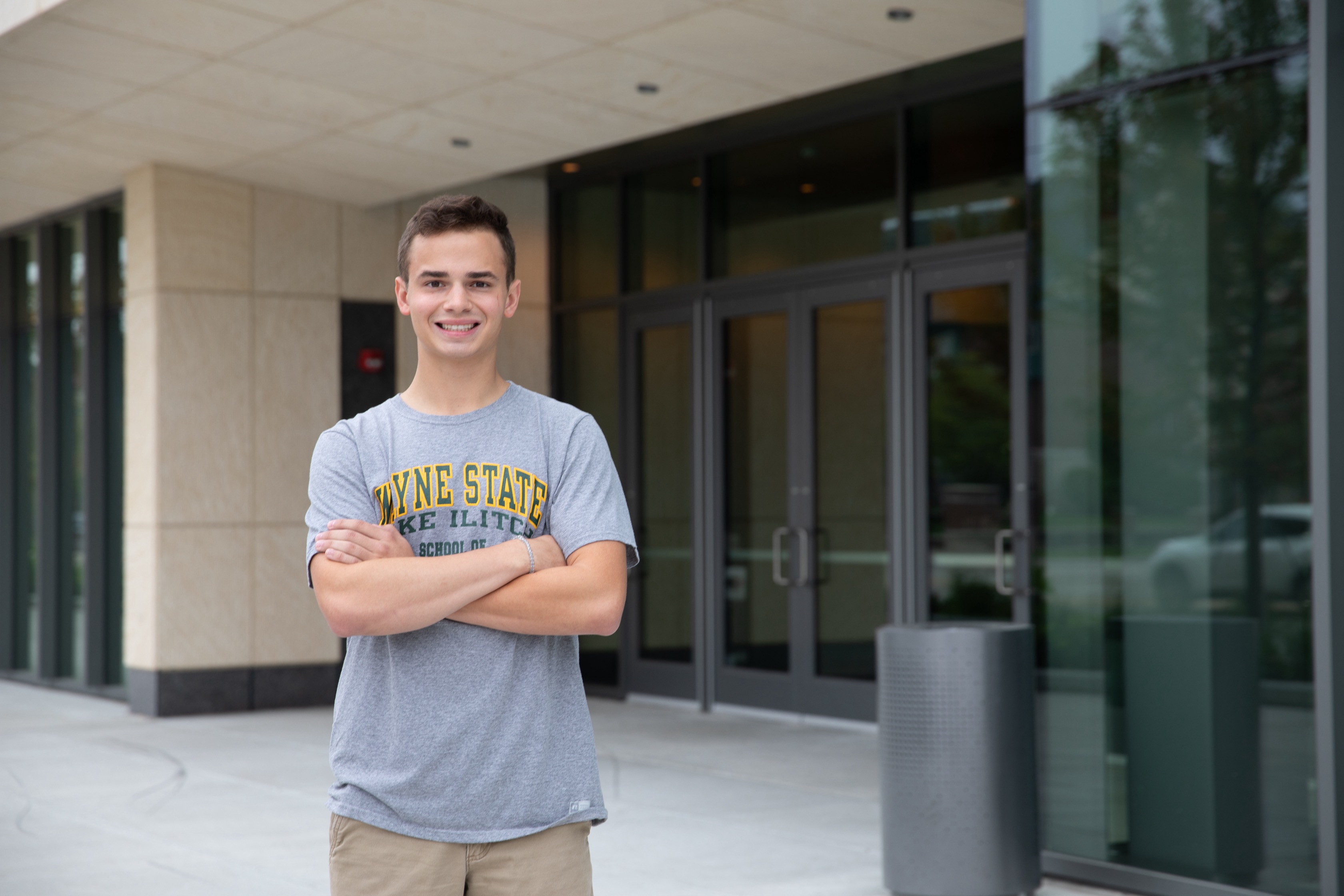 A male student wearing an Ilitch School shirt smiles with his arms folded, standing outside in front of an Ilitch School building