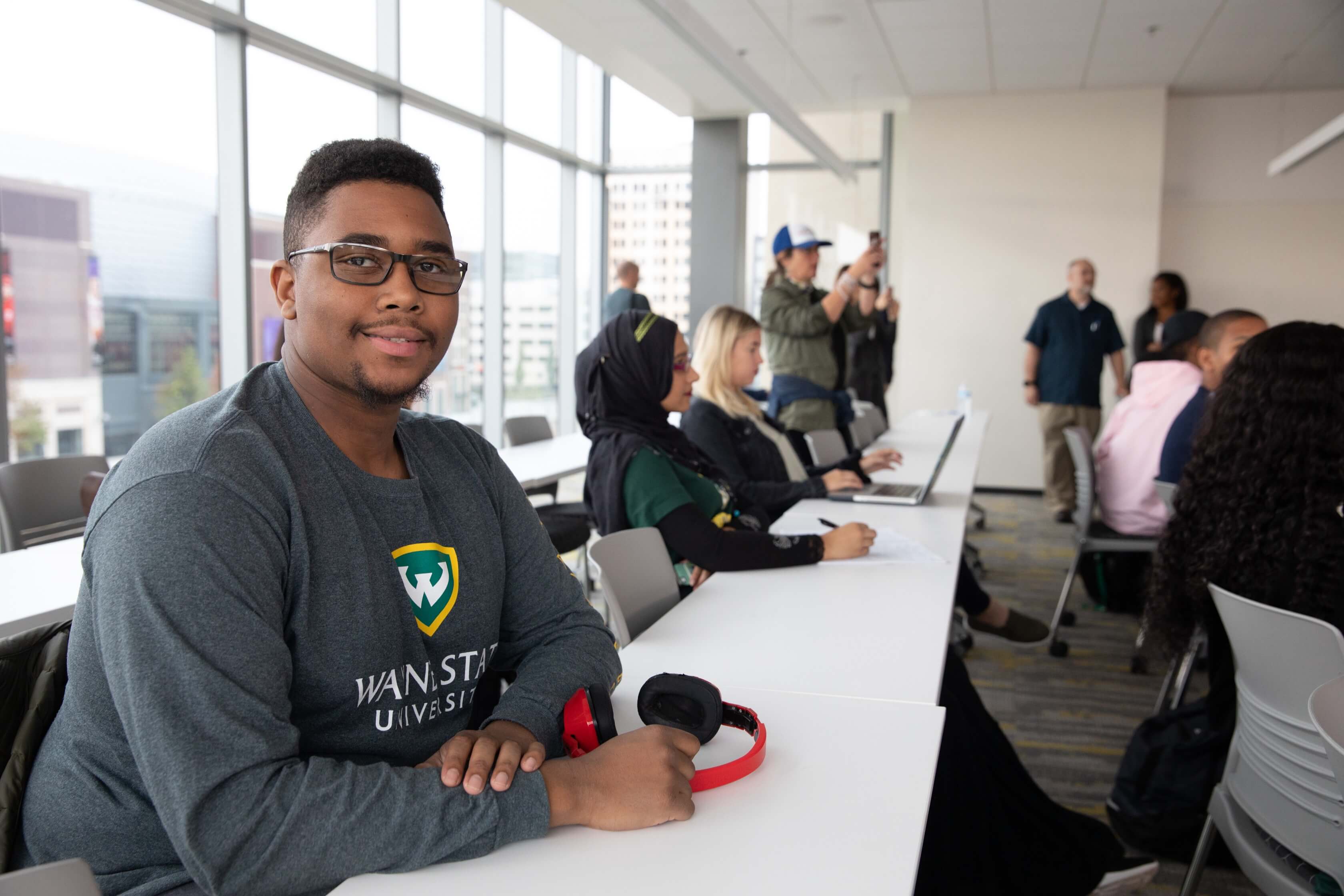 An Ilitch School student wearing a WSU shirt sits at a table in a classroom with headphones on the desk beside him, smiling