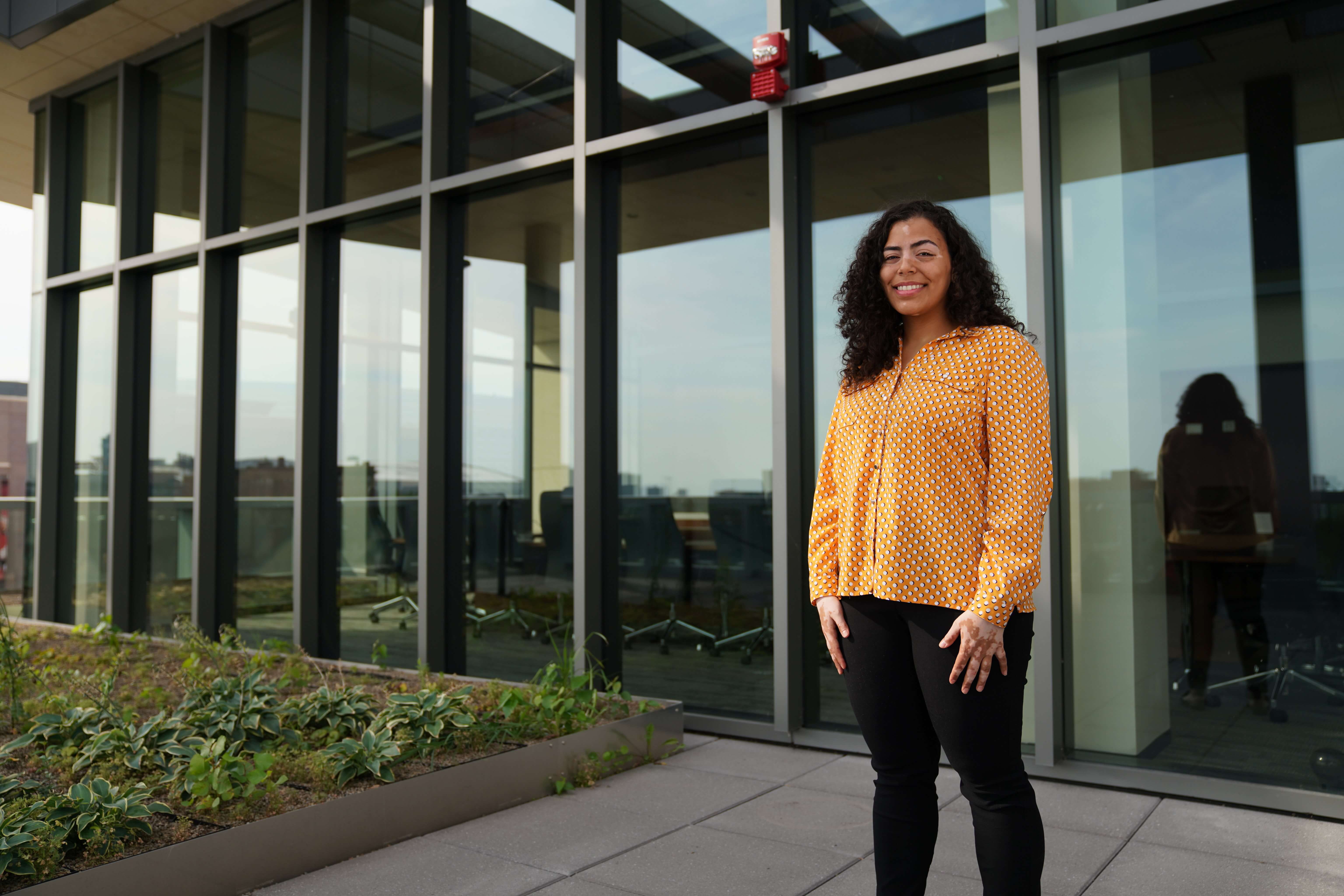 A female business student wearing an orange shirt poses on Ilitch School&#039;s terrace in front of windows