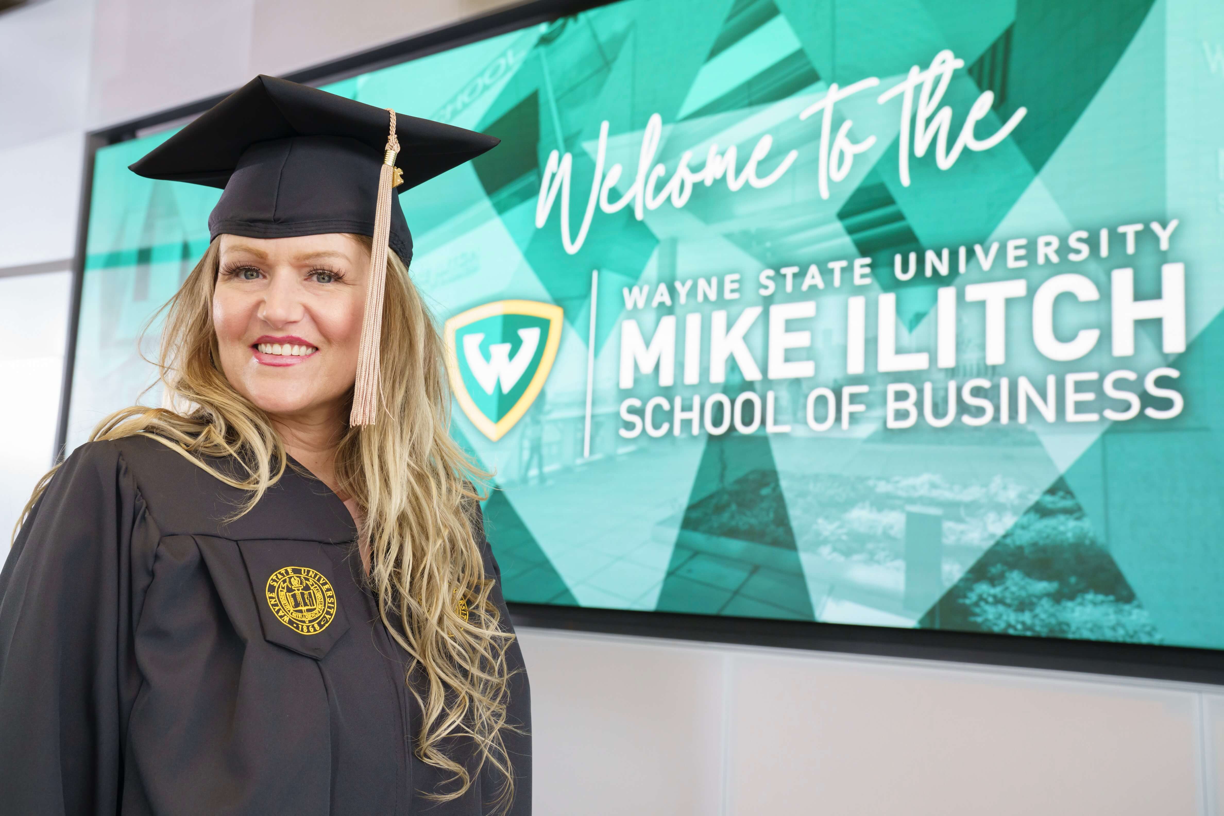 A female business graduate stands inside smiling in her cap and gown in front of a Wayne State Ilitch School welcome sign