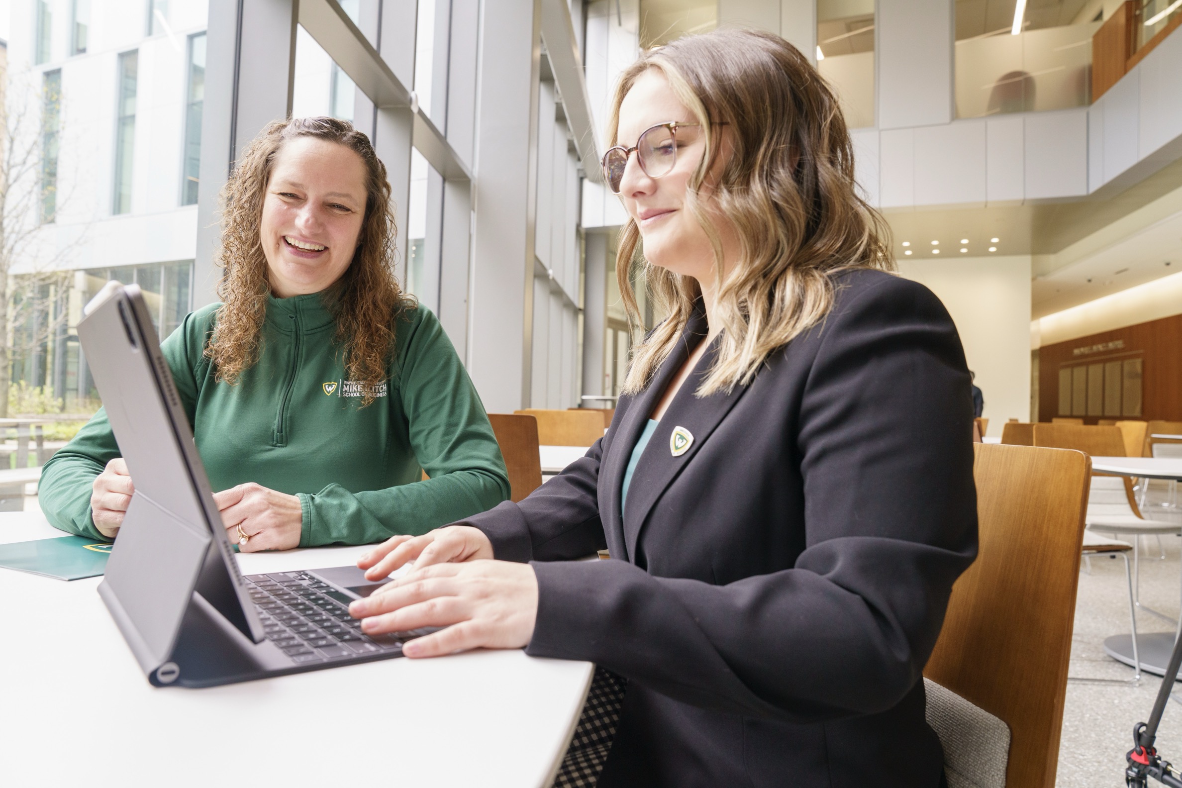 A female undergraduate business student sits at a table with an Ilitch School staff member, smiling and typing on her laptop  