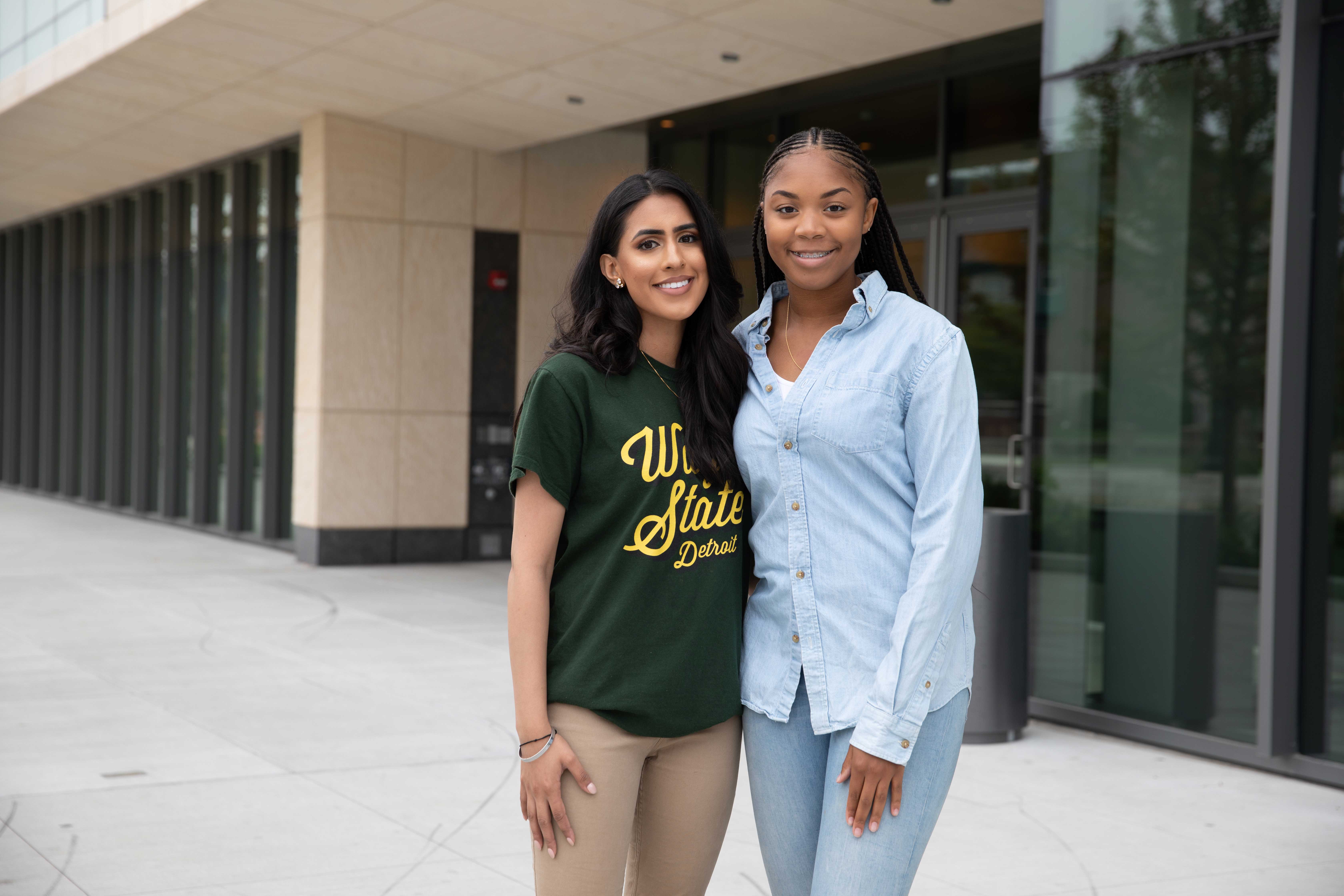 Two female business students pose with their arms around each other in front of an Ilitch School building