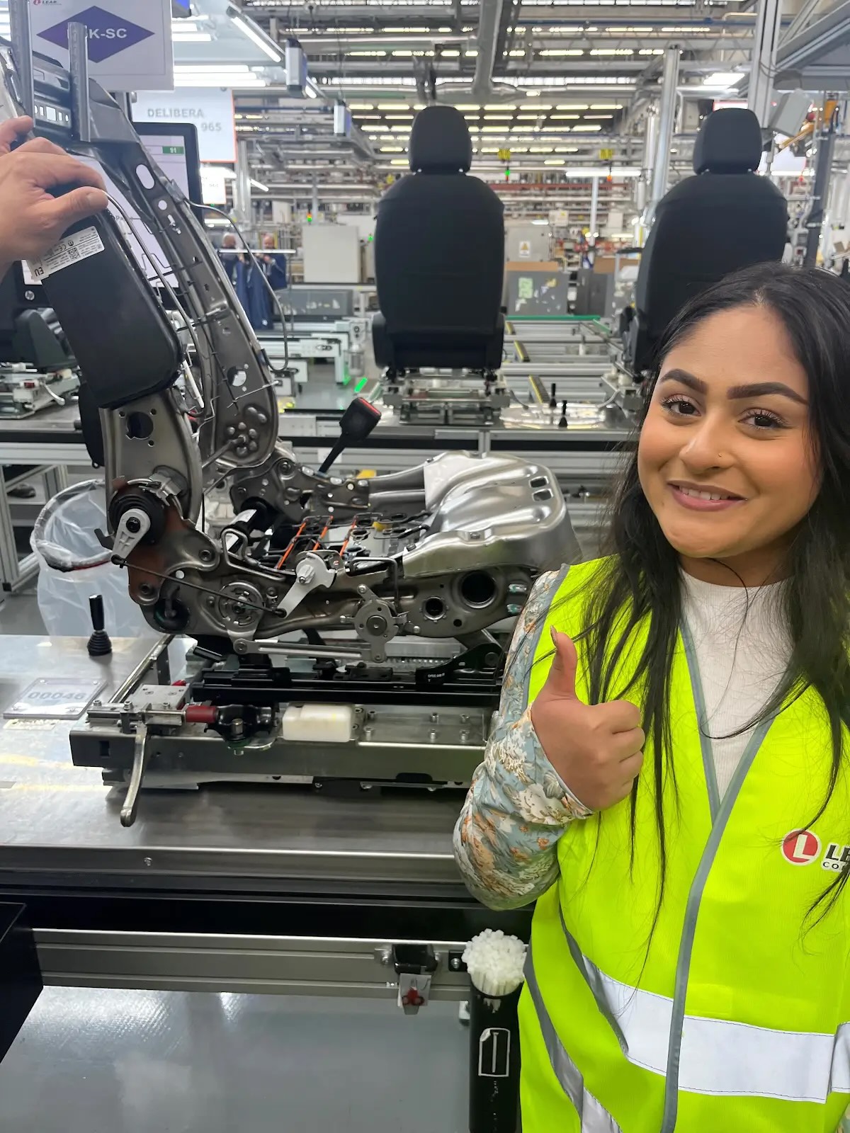 A female business student studying abroad smiles and poses with her thumb up in front of a chair model at Lear Corporation