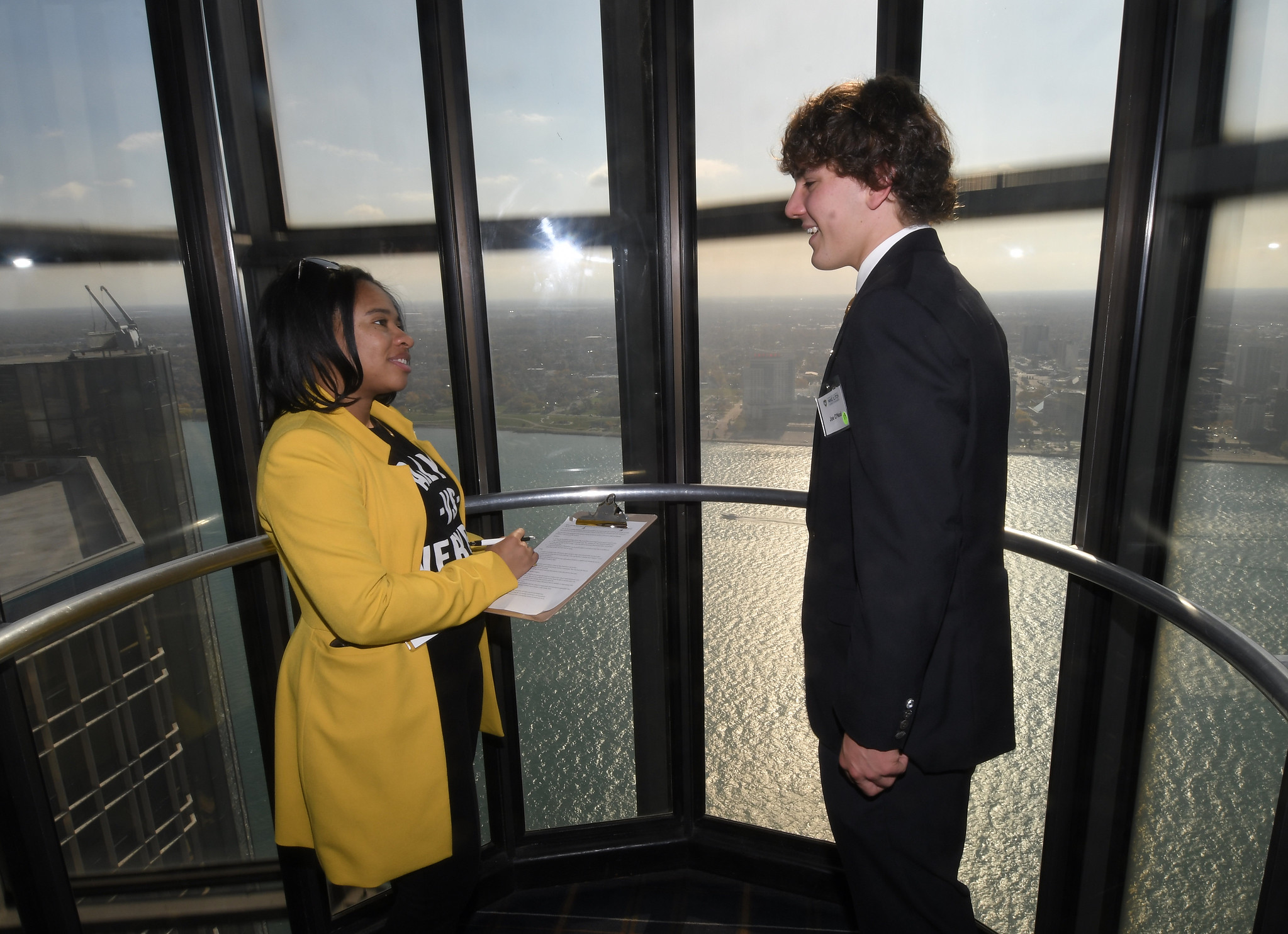 A professionally dressed male business student and female holding a clipboard stand in an elevator overlooking a body of water