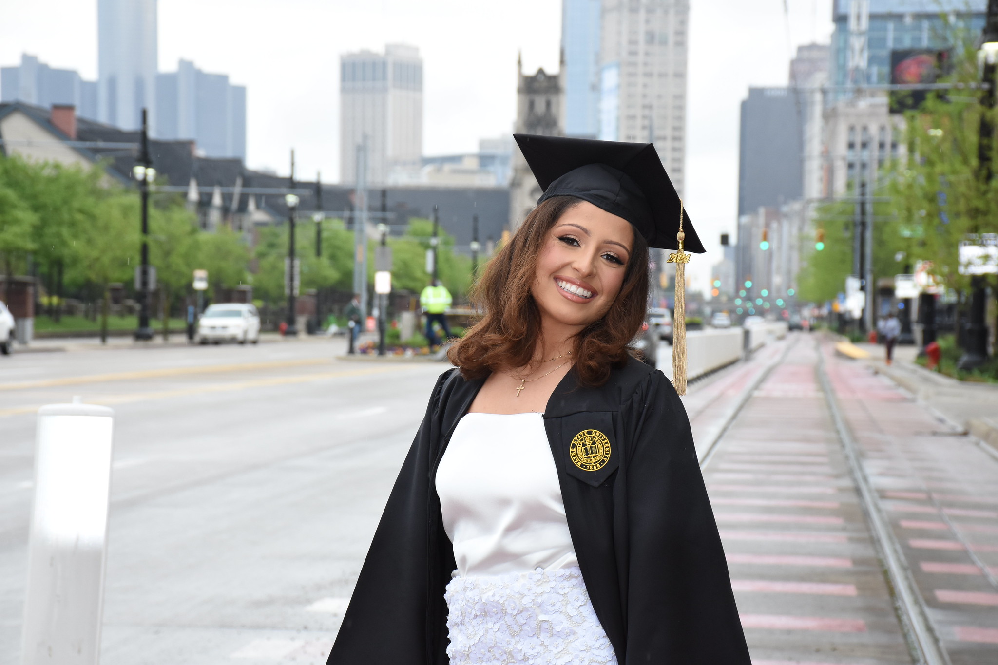 An Ilitch School of Business alumna smiles in a her graduation cap and gown outside of the Ilitch School on Woodward Ave. with the downtown skyline behind her