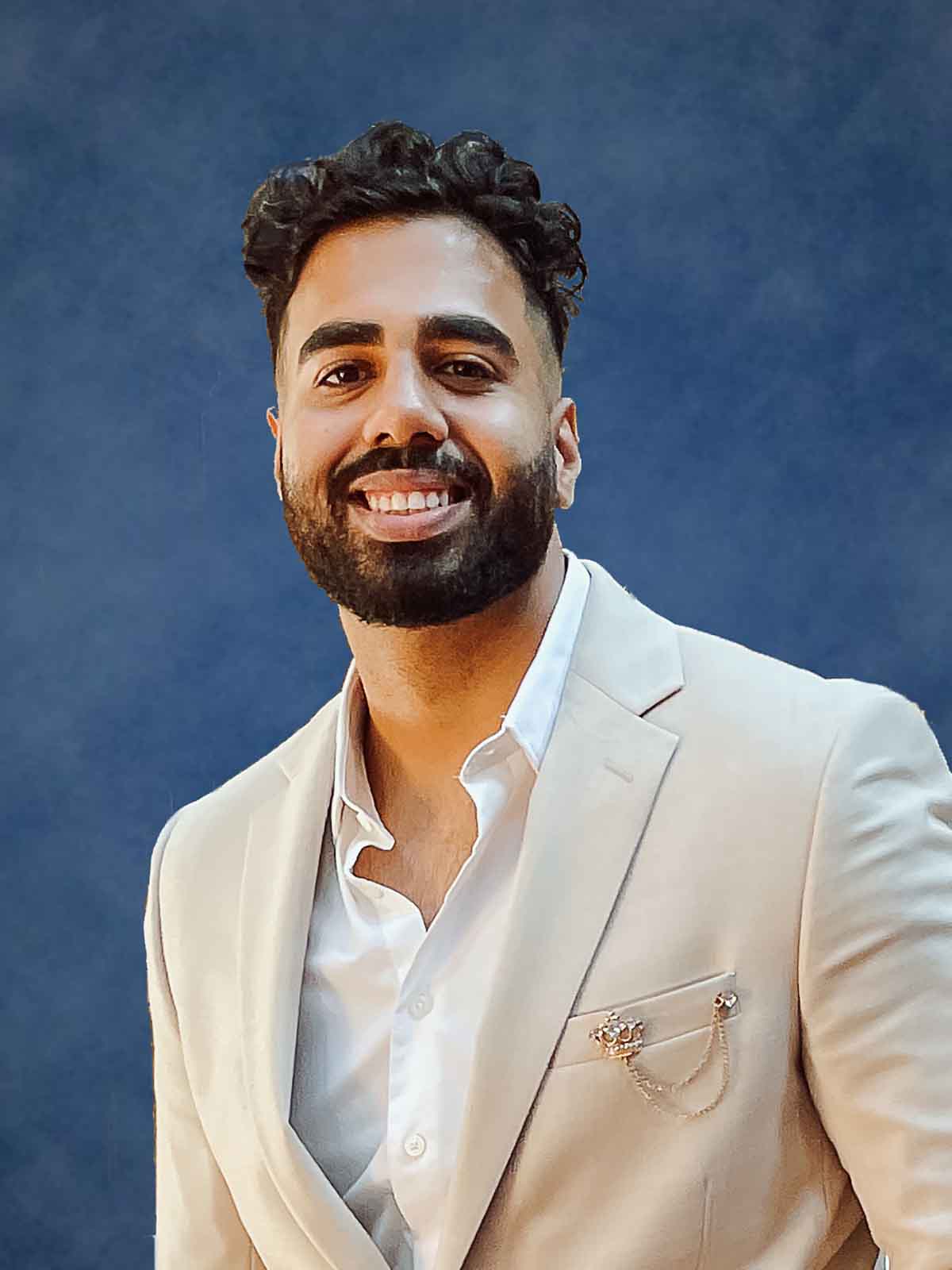 2018 and 2023 Ilitch School graduate, Joy Gope, smiles in a suit in front a blue backdrop for an indoor headshot